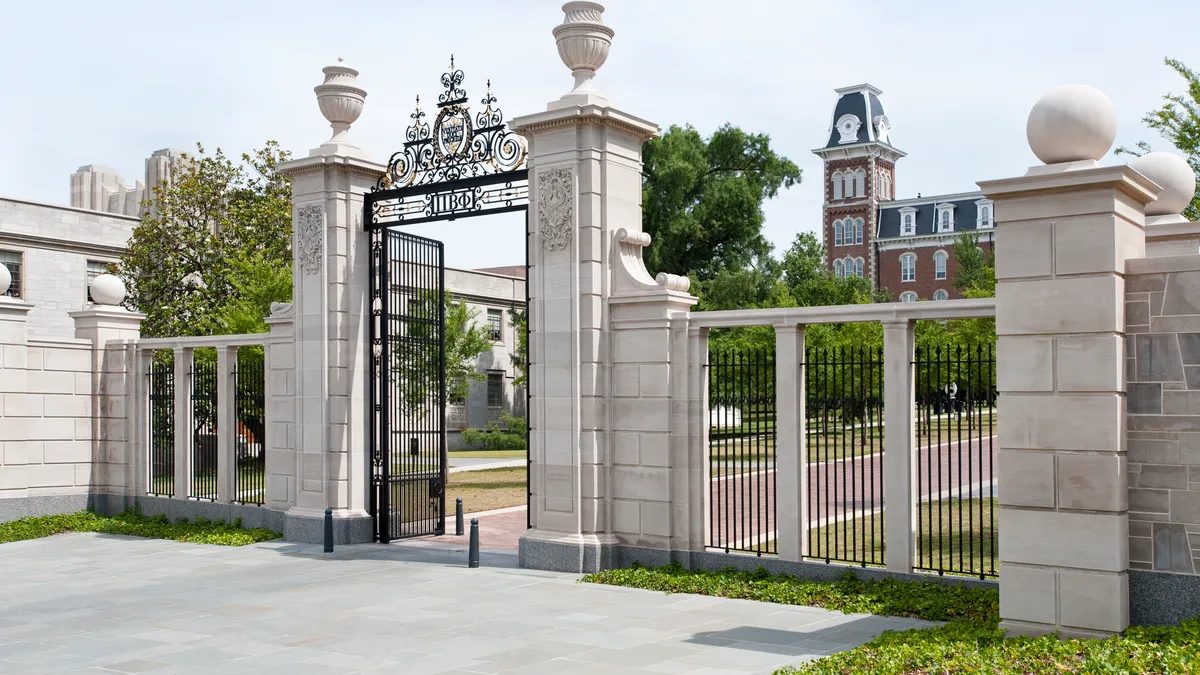 A white stone and wrought iron gate stands in front of classic brick academic buildings.