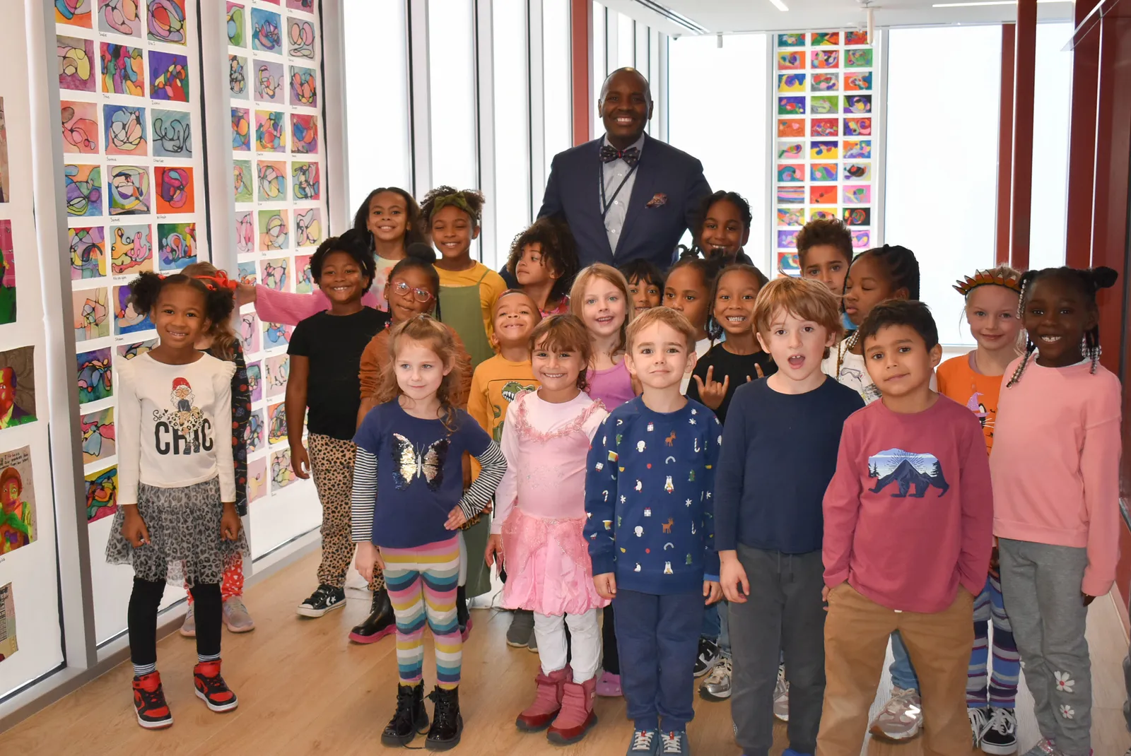 Young students stand in front of and around an adult in a school hallway. Everyone is looking at the camera. The walls of the hallway show student artwork.