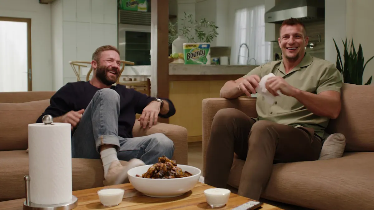 Rob Gronkowski and Julian Edelman sit on a couch near a bowl of wings and paper towels.