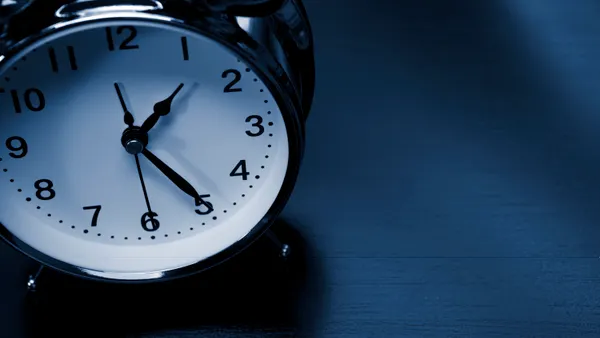 An alarm clock with the hands pointing to 1:25 sits on a table.