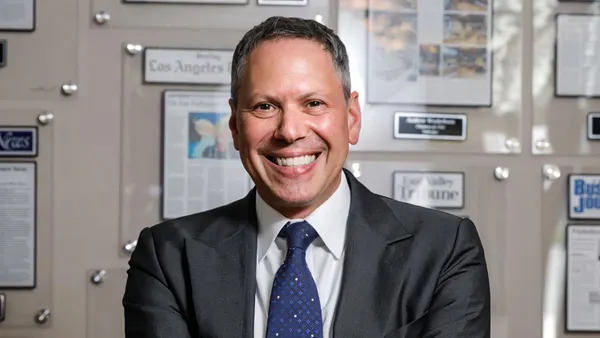 An image of a person wearing a suit with arms crossed and smiling. The person is in front of a wall with news clips in the background.