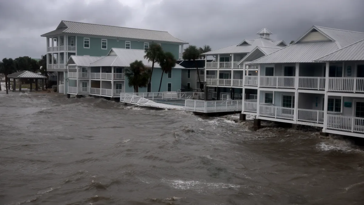 Ocean water comes up against houses and palm trees. The sky is stormy and cloudy.