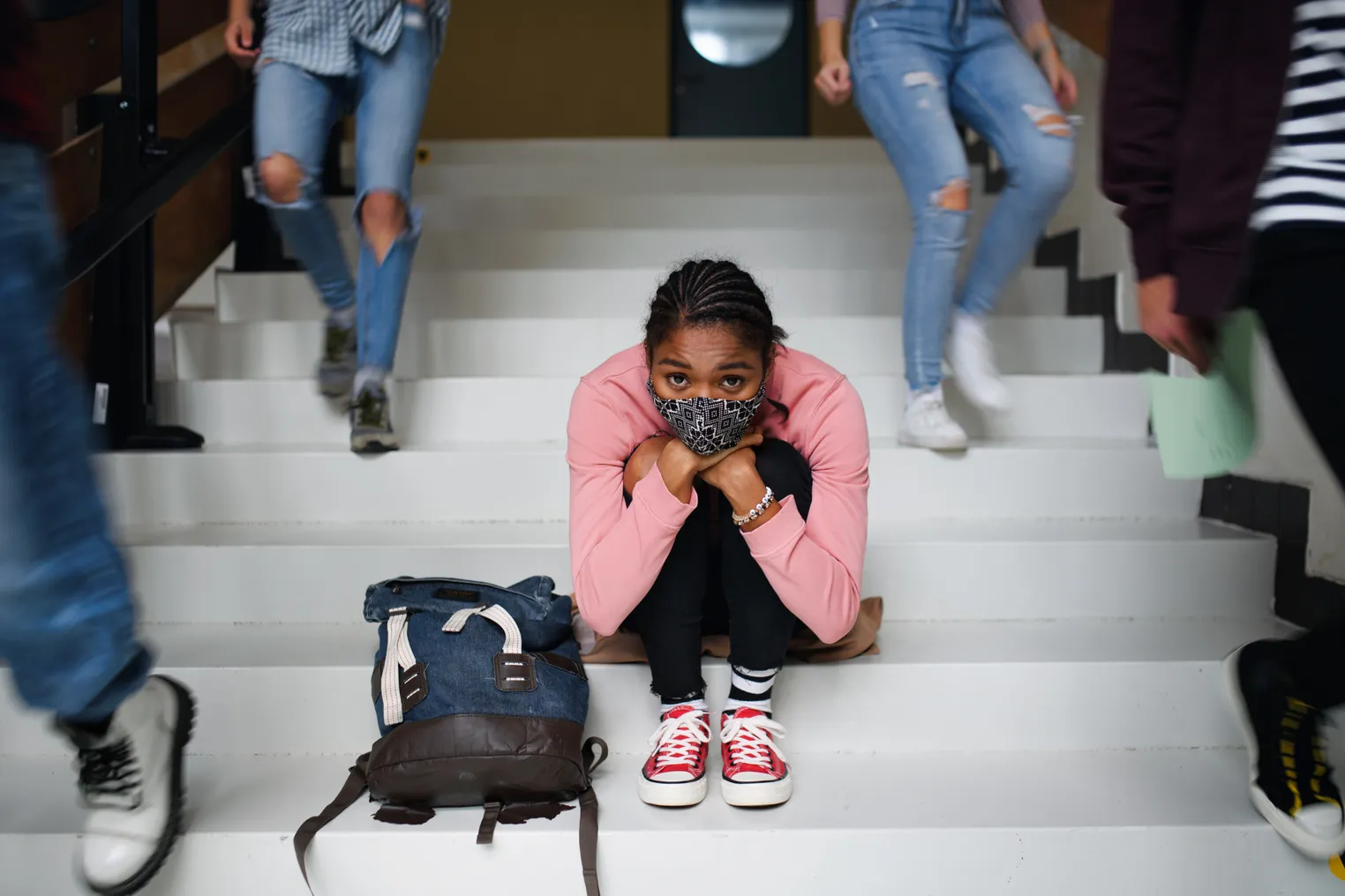 A student wearing a face mask sits on the steps in a school building staring ahead as other students walk around.