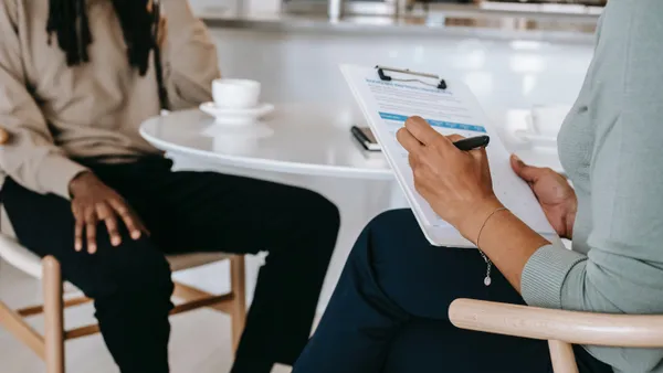 A Black person with dreadlocks sits across from a person writing on a clipboard