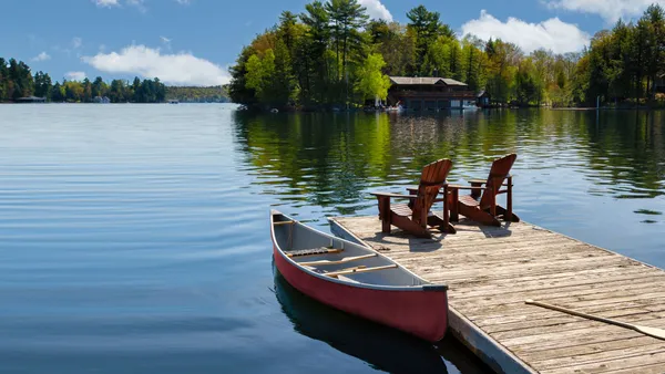 Two Adirondack chairs on a wooden dock overlook a lake. Nearby, a red canoe is tied to the pier, alongside life jackets and a paddle.