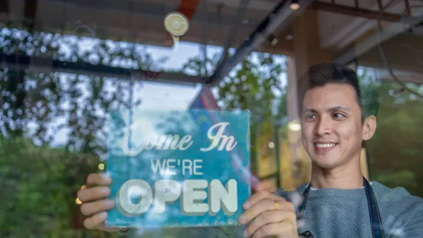 A photograph of a person behind class touching a blue sign that says "Come In We're Open."