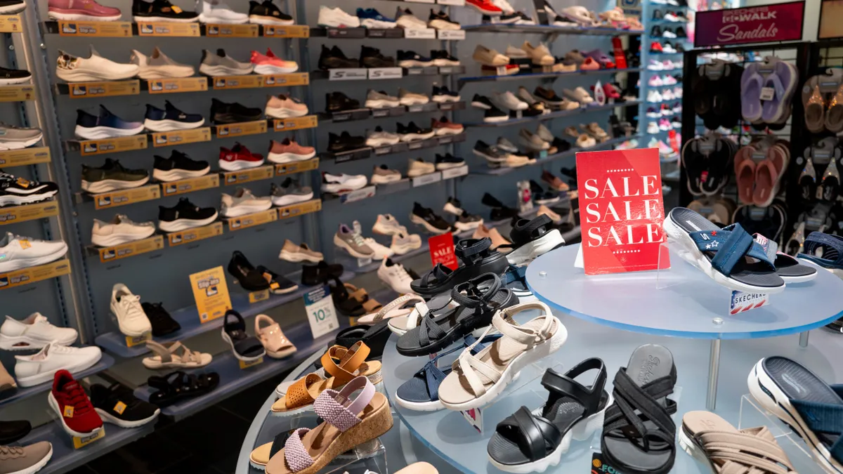 A wall of shoes on display in a Skechers store. A table of shoes with a sale sign on it stands in the foreground.