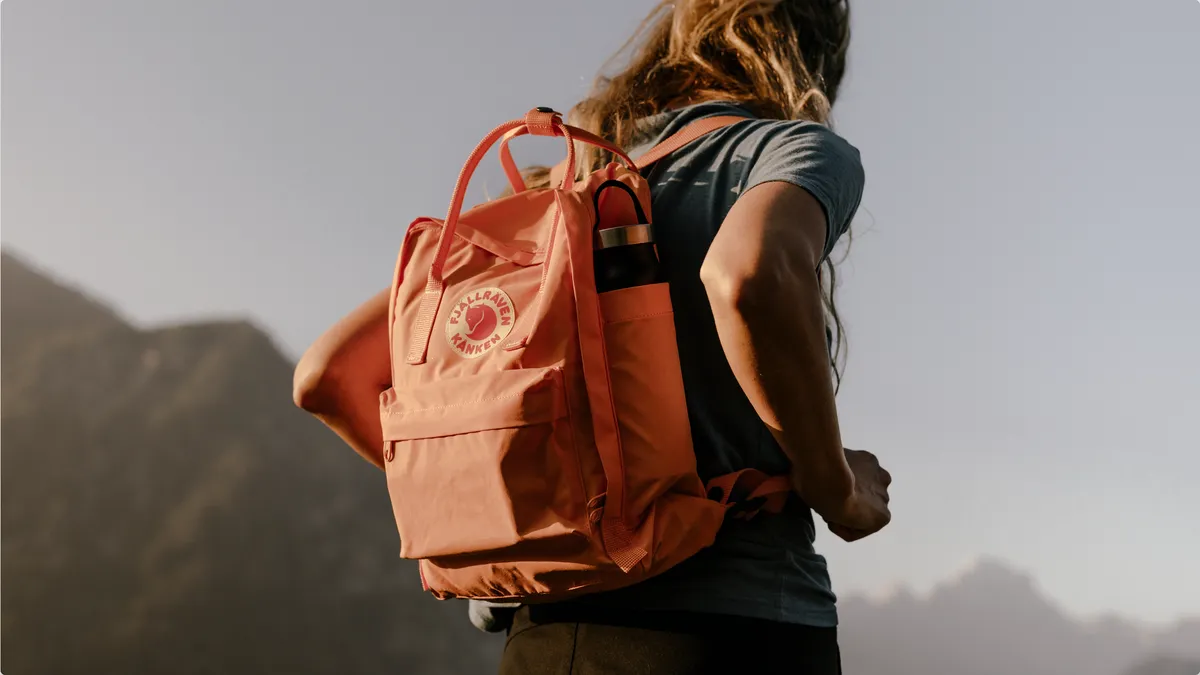 A person carrying an orange Fjällräven backpack with a water bottle on the side.