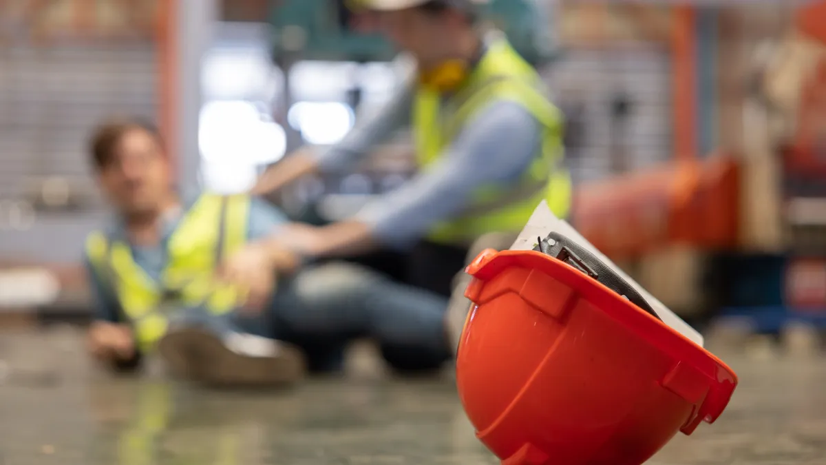 A close-up of a red hard hat with a blurred injured factory worker and another helping them wearing yellow vests in the background.