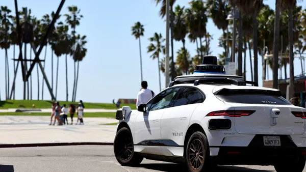 A white car labeled "Waymo" drives among palm trees and people.