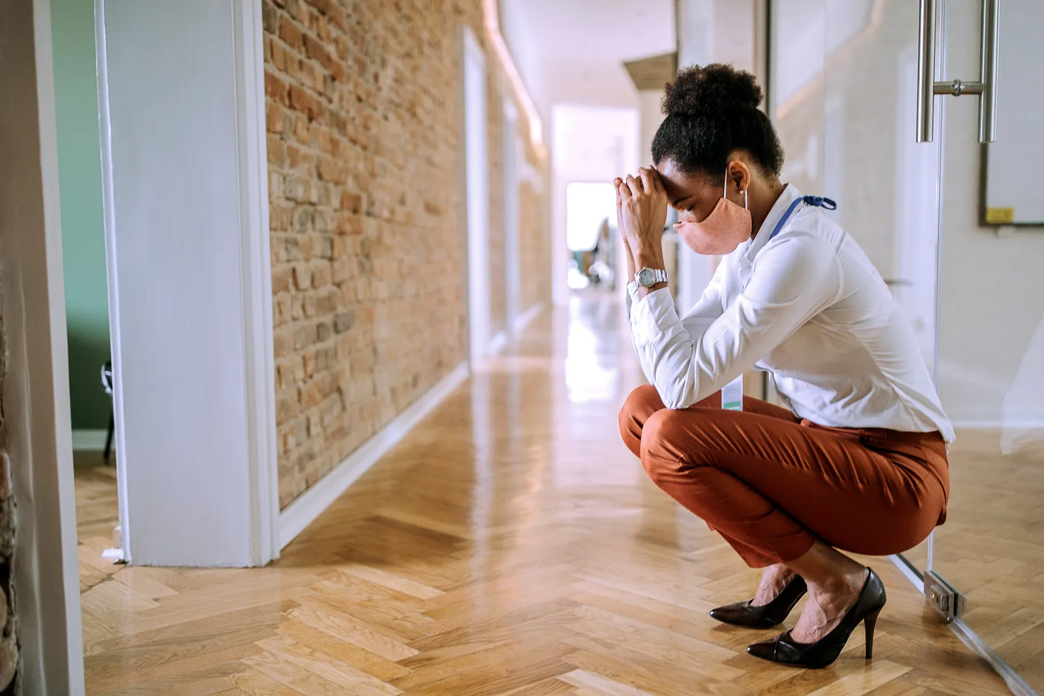 A masked Black person squats in distress in their workplace hallway