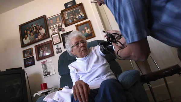 A person sits in a chair in while another person attaches a blood pressure cuff on their arm. In the background is a wall with several framed photos.