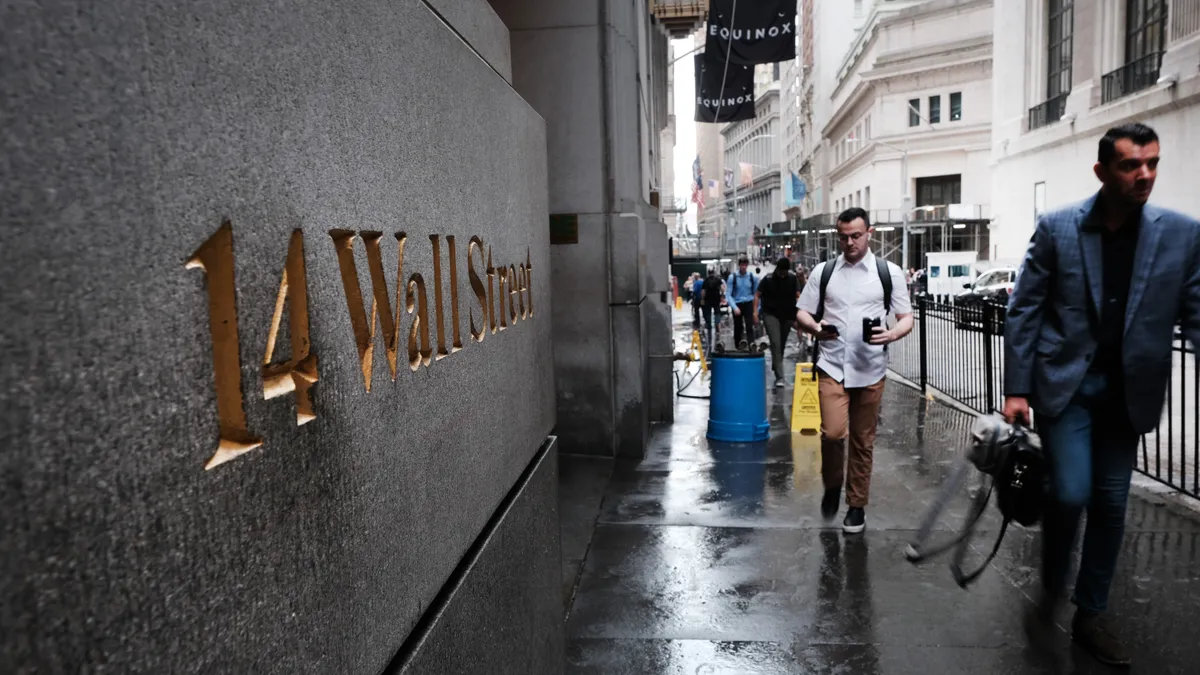 Pedestrians walk beside a stone wall with gilded letters spelling out "14 Wall Street."