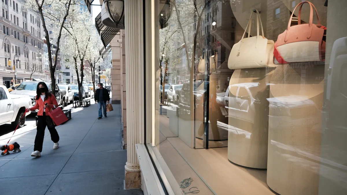 People walk on a sidewalk in front of a luxury store display showcasing handbags.