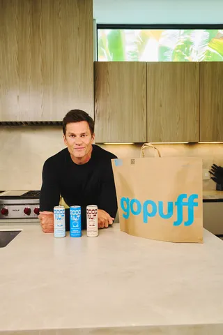 Tom Brady next to a counter top that has a Gopuff bag and three cans of Gopuff coconut water on it.