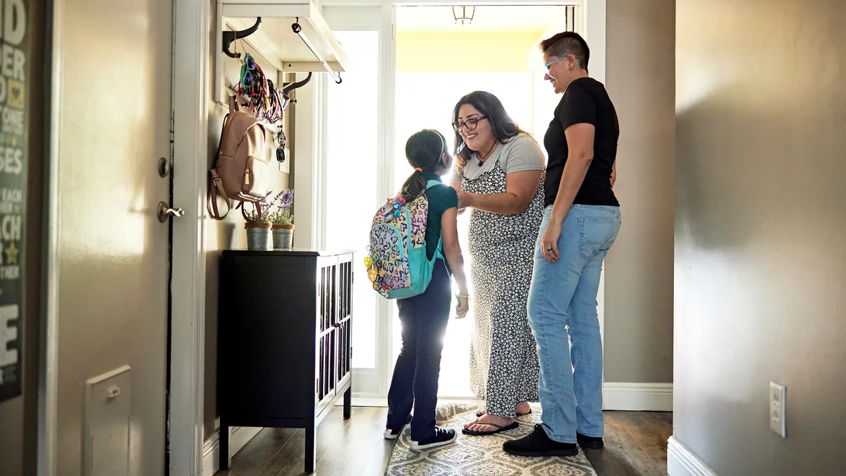 Full length view of smiling women standing at sunny entrance to family home and helping 8 year old girl with backpack before leaving.