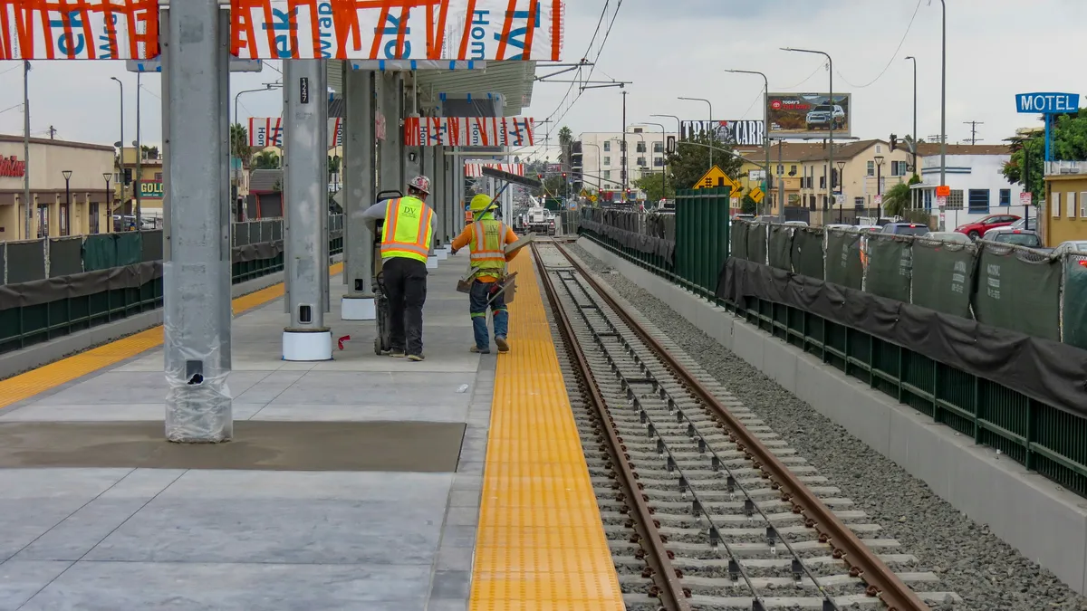 A light rail station under construction in Los Angeles is seen looking along a platform with tracks on either side and two workers in yellow vests.