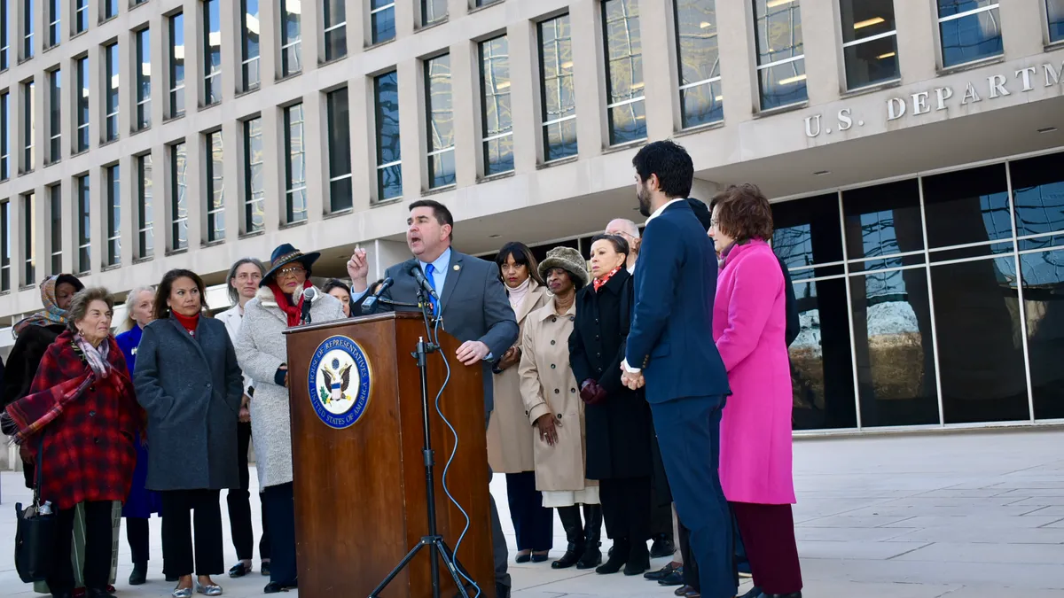 A group of people are standing outside in front of the U.S. Department of Education in Washington, D.C. They are standing behind a podium where one person is speaking.