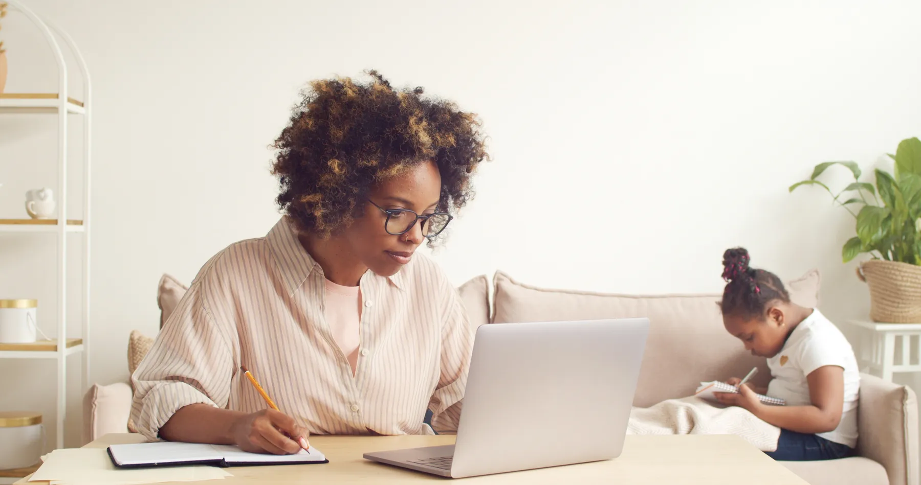 A woman works at her laptop, while a child sits on a couch in the background.