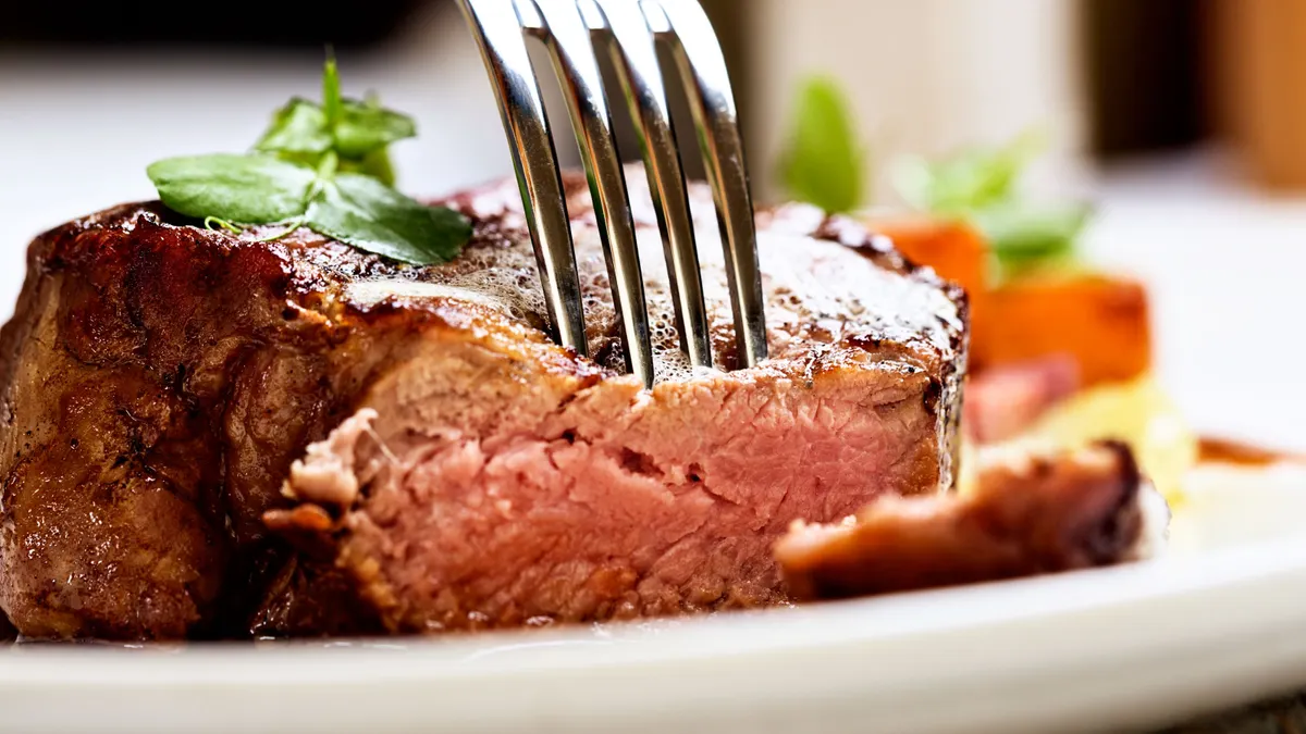 An image of a fork piercing a steak that sits on a white plate.