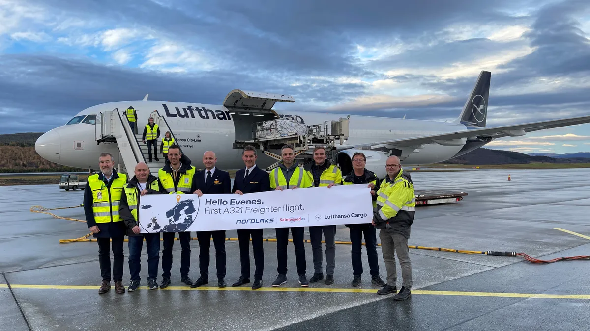 A team of people hold a sign that welcomes the first freighter flight to Evenes with a freighter aircraft in the background.