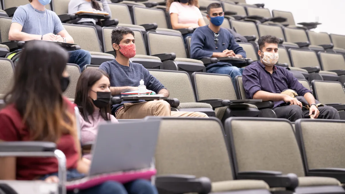 A dozen masked young people sit scattered across a lecture hall.