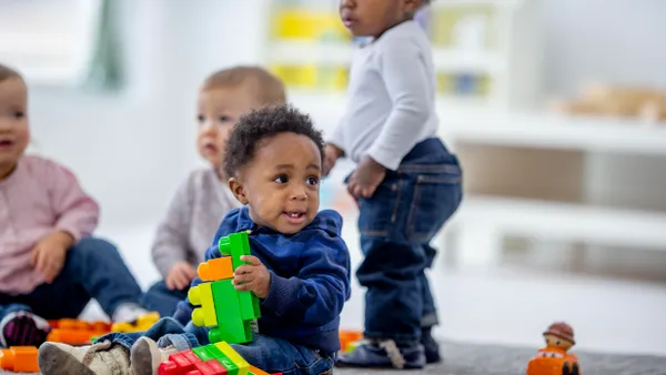 Three toddlers are pictured sitting on the ground, while one is standing. The toddler in the center is looking off to the side and is holding a plastic toy.