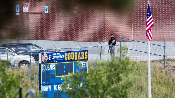A police officer with a long gun is seen in the distance near the exterior of a red brick school building. In the foreground is an athletic scoreboard that says the school name.