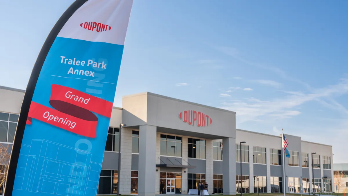 A tan building with the red DuPont logo above the glass entrance doors in the center, and a blue grand opening flag on the left.