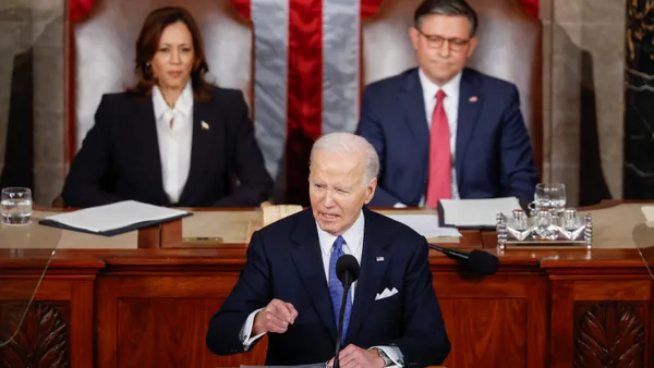 President Joe Biden speaks at a podium with Vice President Kamala Harris and Speaker of the House Mike Johnson behind him.