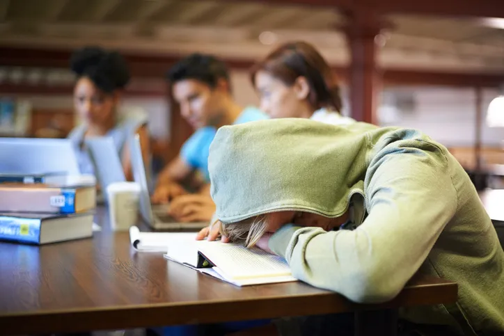 A student sitting at a desk is shown with their head down on a desk and their hood pulled over their head. Other students can be seen working in the background.