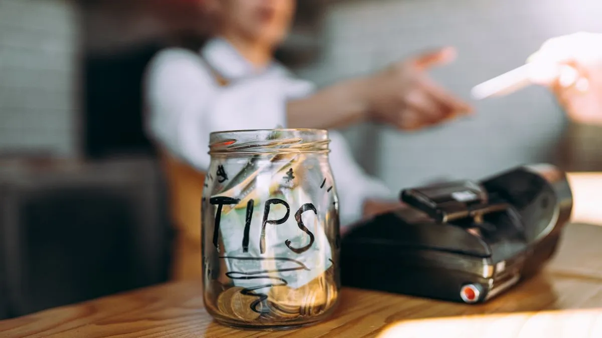 An image of a tip jar in the foreground with a customer paying by credit card in the background