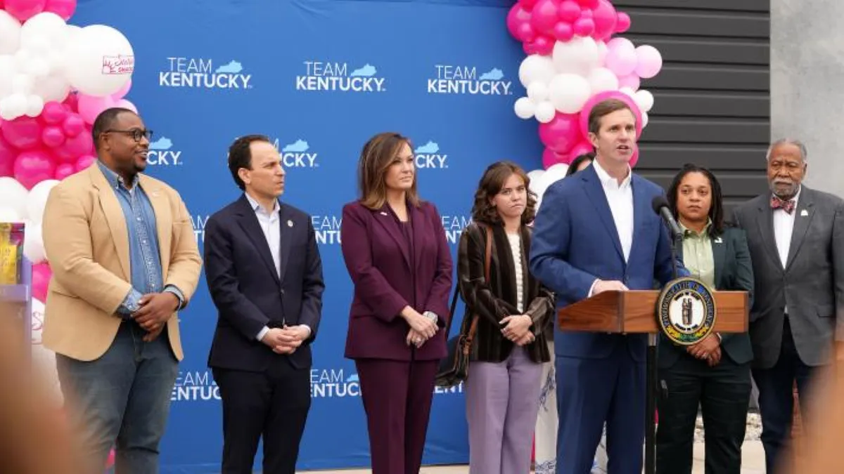 People lined horizontally behind a person in a blue suit with a podium and microphone in front.