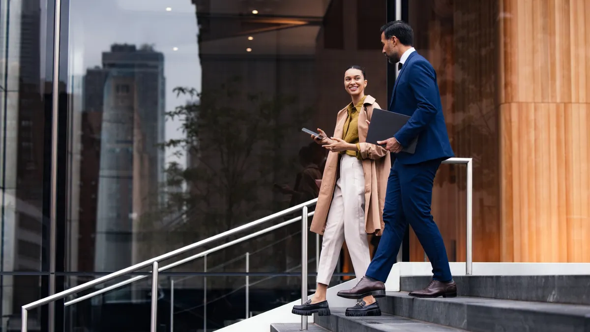 A man in a blue suit carries a laptop while walking down stairs with a smiling woman holding a phone.