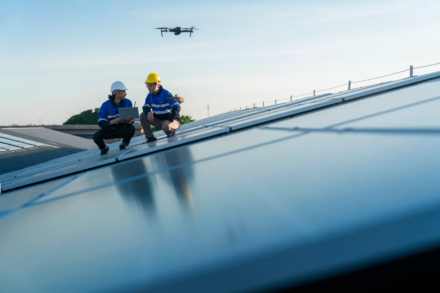 Two technicians work on a solar panel as a drone flies overhead.