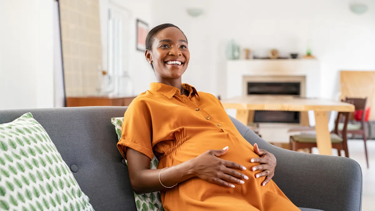 A Black pregnant woman rests in a living room while holding her belly.