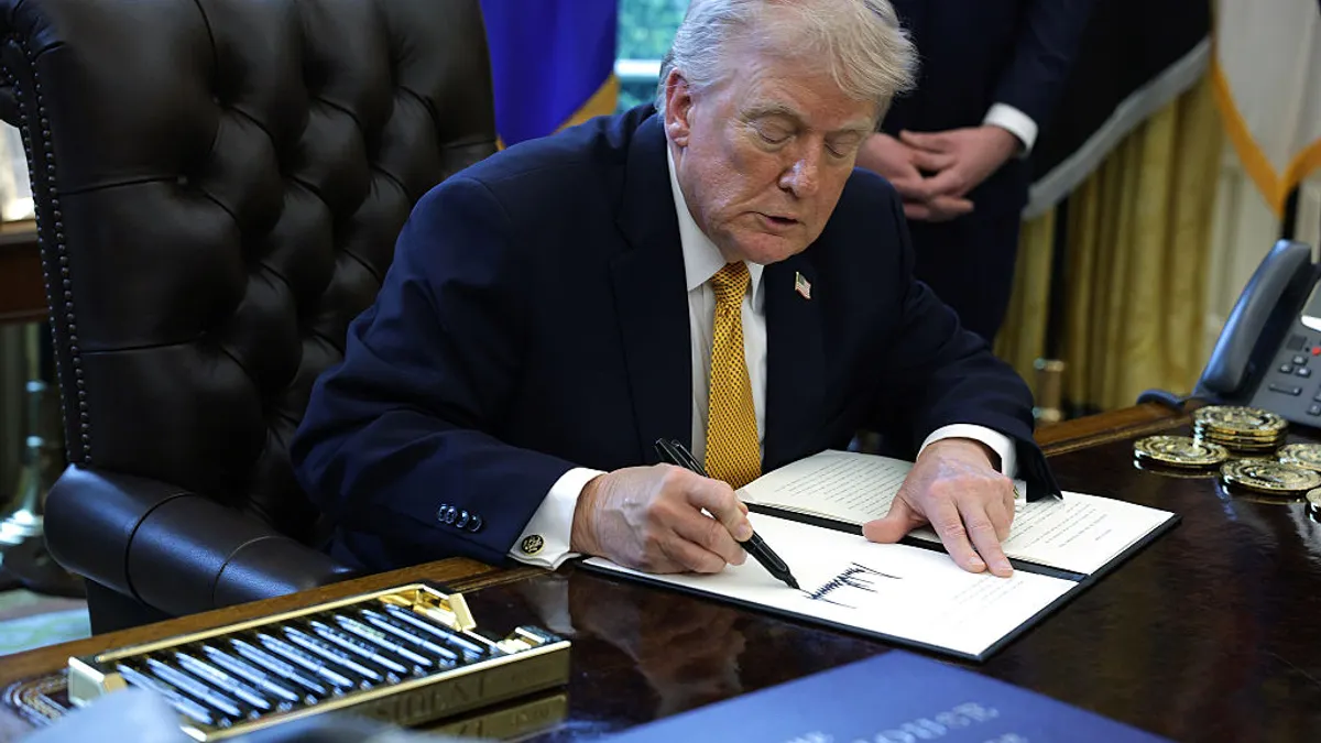 Wearing a suit and gold tie, President Donald Trump leans forward over a desk with a pen and signs an executive order.
