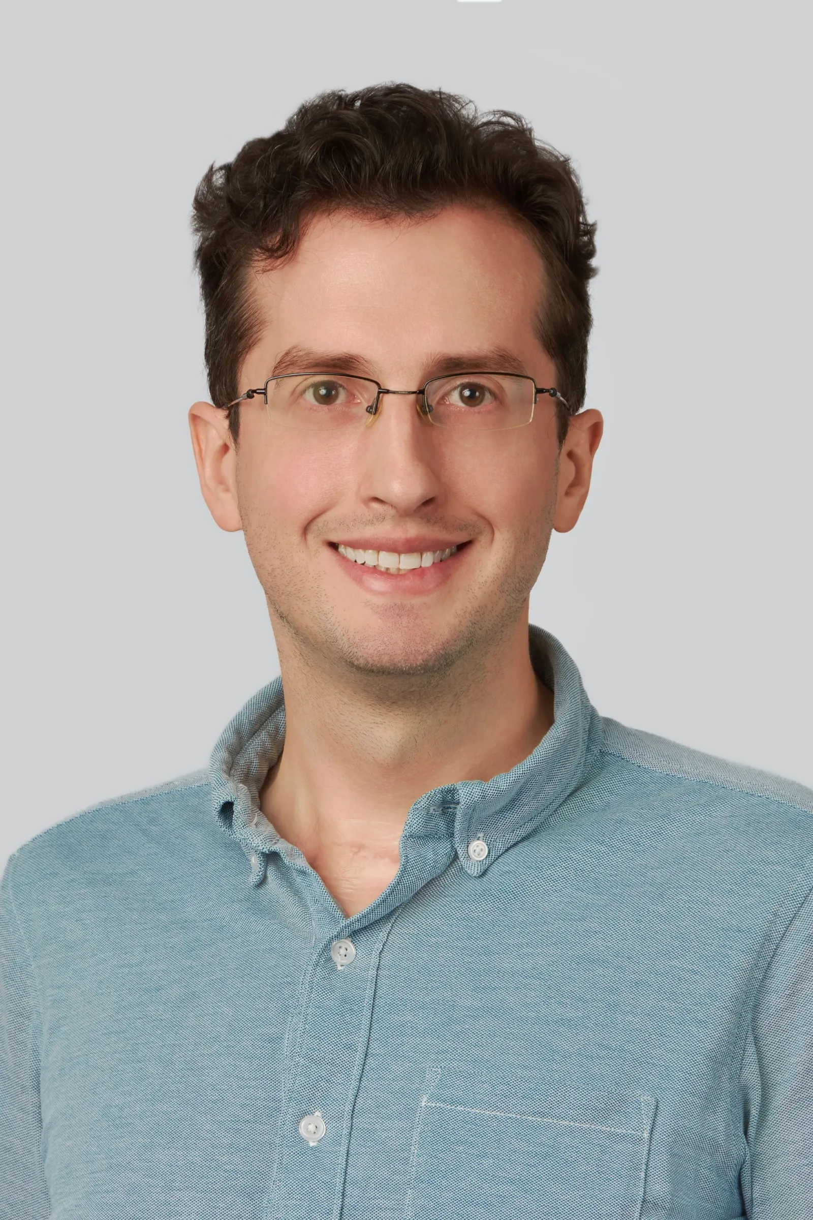 A headshot of a person with short dark hair, wearing eyeglasses and denim-shade button up blue shirt.