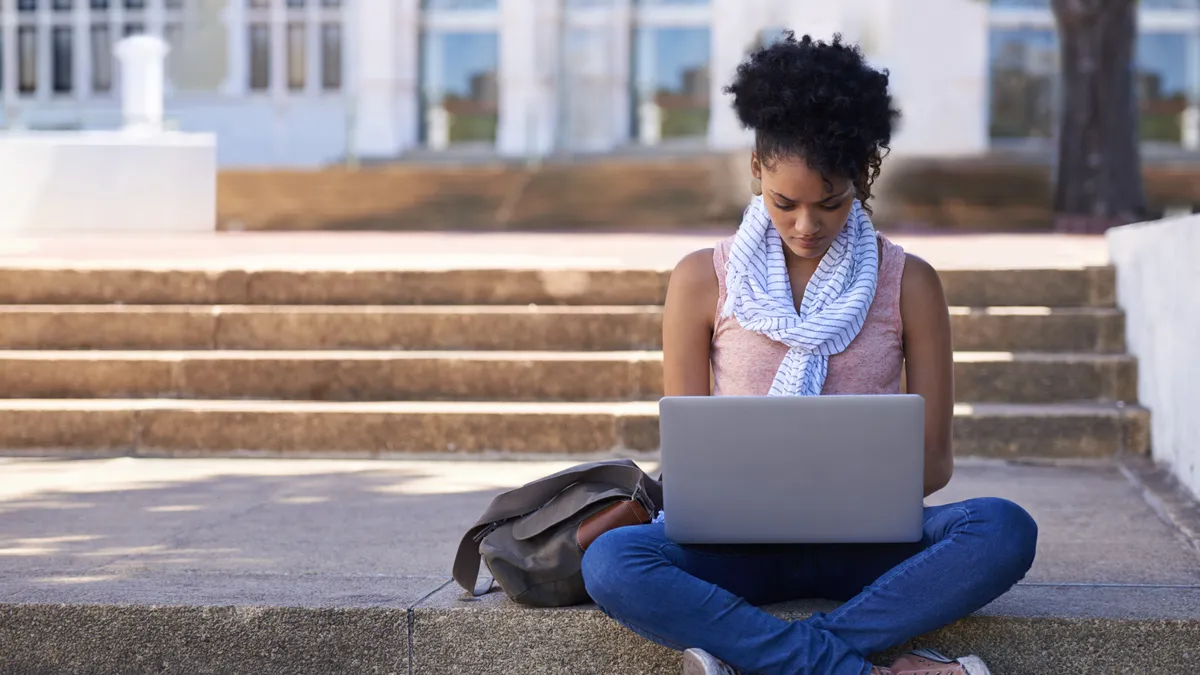 A woman sits on the steps of her college while doing research on her laptop.