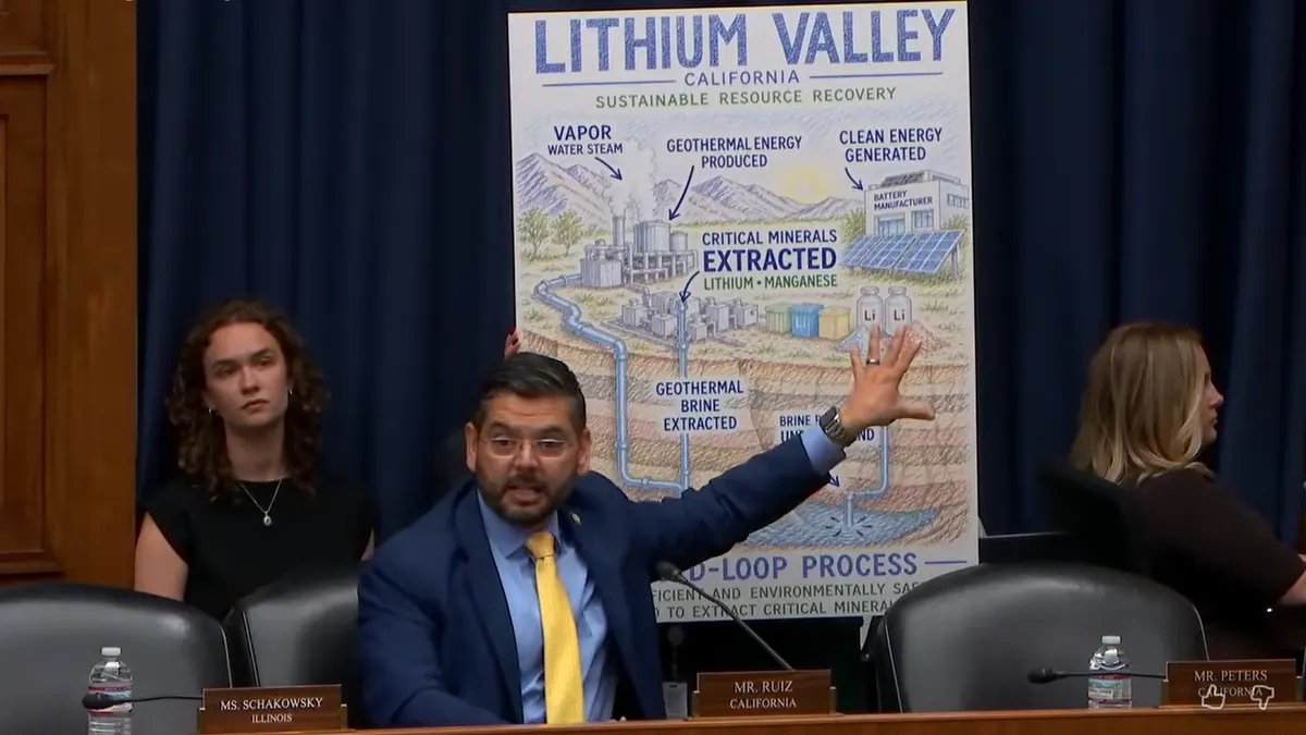 Screenshot of a man in a business suit and glasses waving his left hand at a colorful poster that maps out the critical mineral process in California.