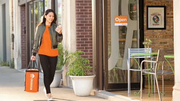 A woman with brown hair is looking at her phone and carries an orange bag with white "Grubhub" logo.