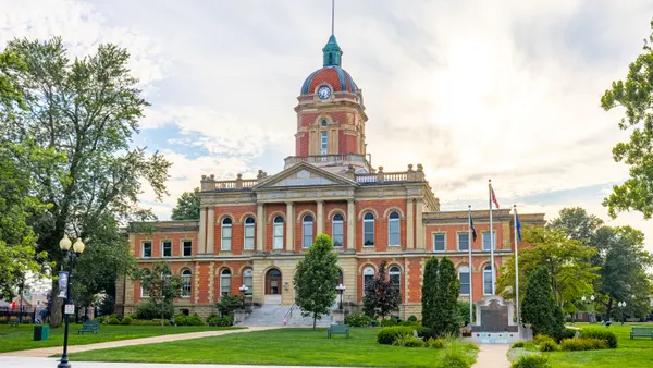 A brick-and-stone government building is surrounded by trees, grass and bushes. Flags and a small monument are shown in the right foreground.