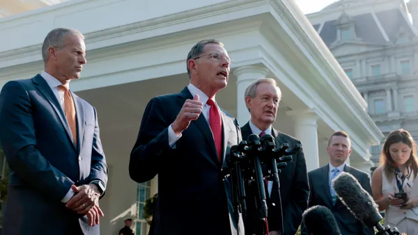 A man wearing a suit and tie speaks into microphones while two men wearing suits and ties look on.