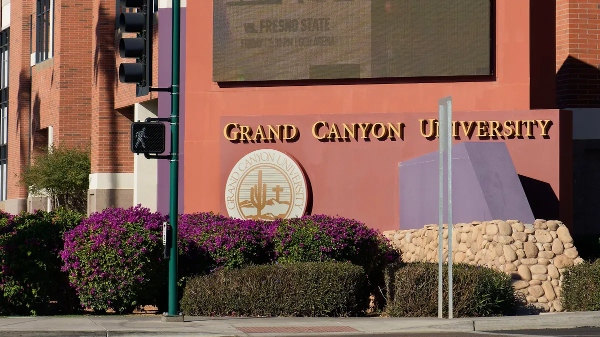 A large "Grand Canyon University" sign is adhered to a red stucco wall; above the sign is a large screen monitor.