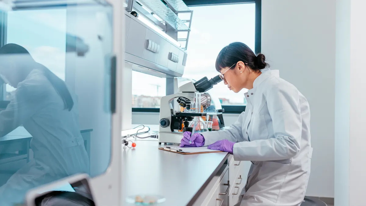 A person in a white lab coat takes notes while looking into a microscope.