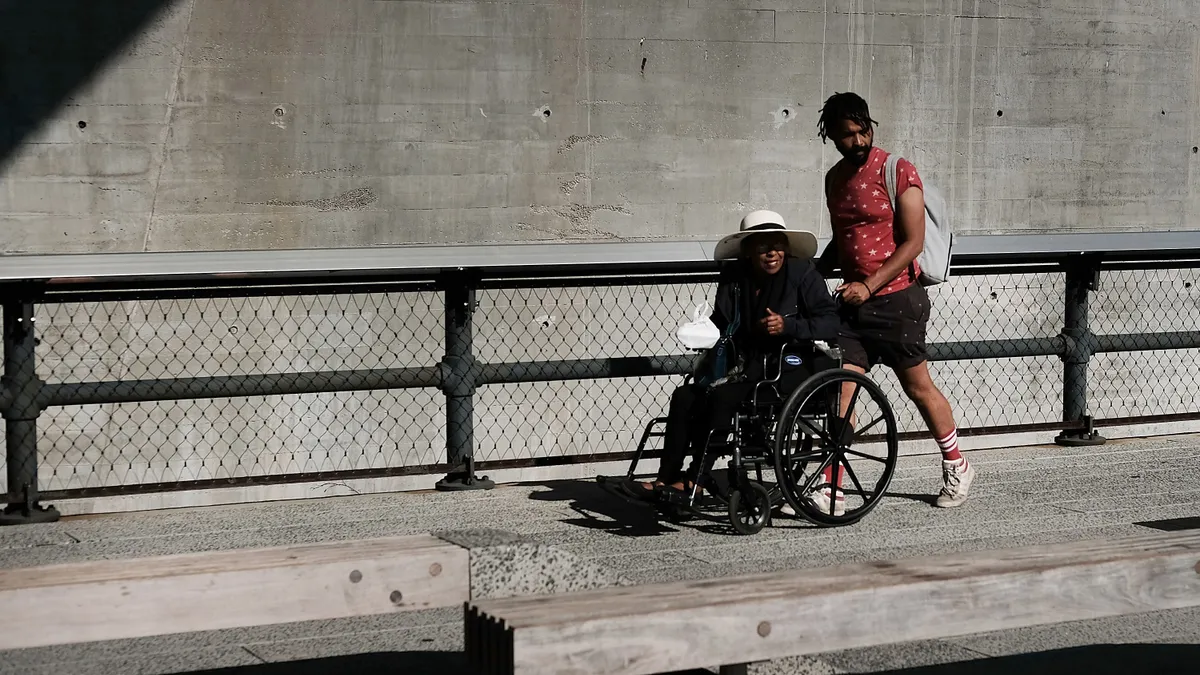 A man in a t-shirt and shorts pushes a person wearing a white hat in a wheelchair along a concrete walkway beside a fence with a wall behind.