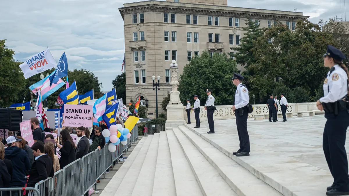 A group of protestors stand behind metal barricades while police stand parallel to the crowd.
