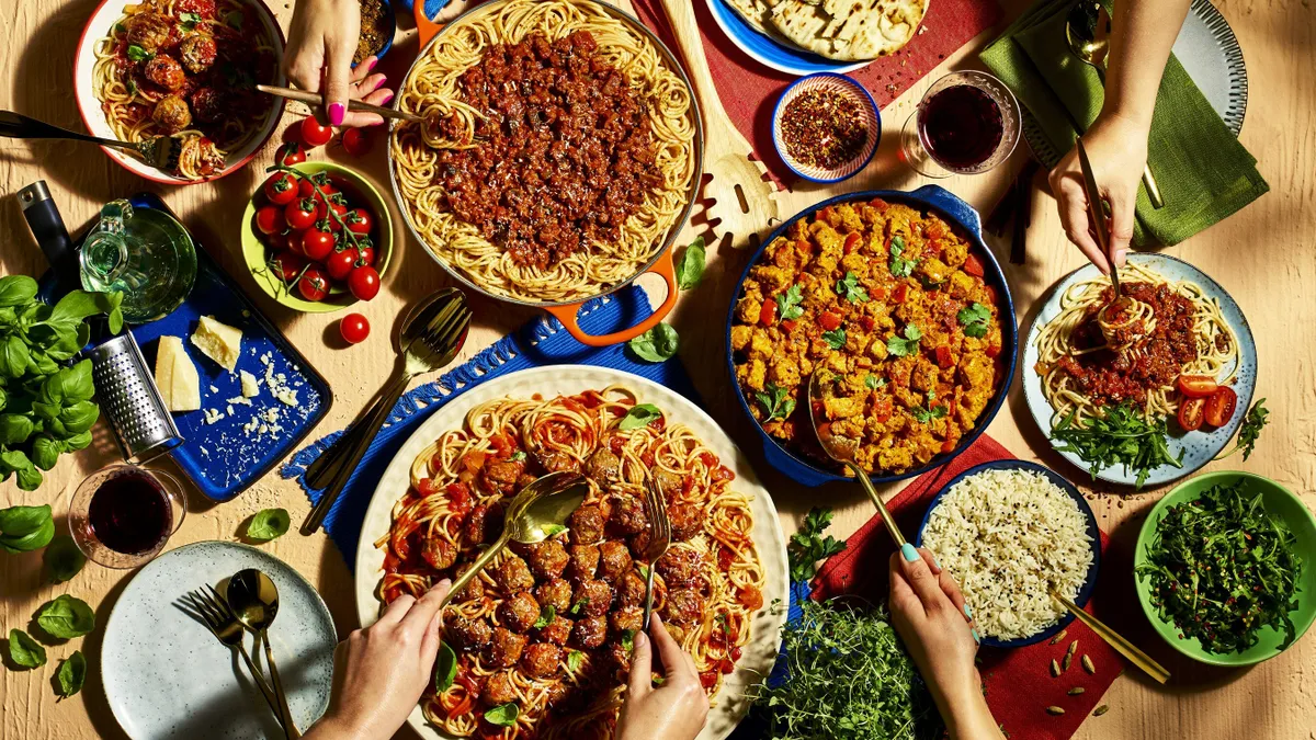 Dishes of food including spaghetti and meatballs, meat curry, pasta with meat sauce, rice and frisee salad fill a table. Four hands with utensils are scooping up some of the food.