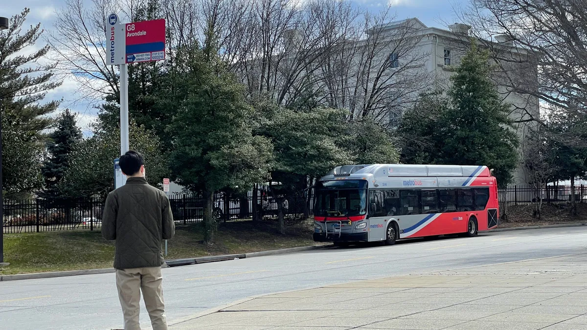 A man standing at a bus stop while a silver and orange bus is stopped across the street in front of trees and a white buildng.