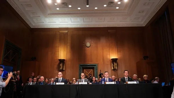 Four people sit at a long table with microphones in front of them, with a crowd of people seated behind them, in a Congressional building.